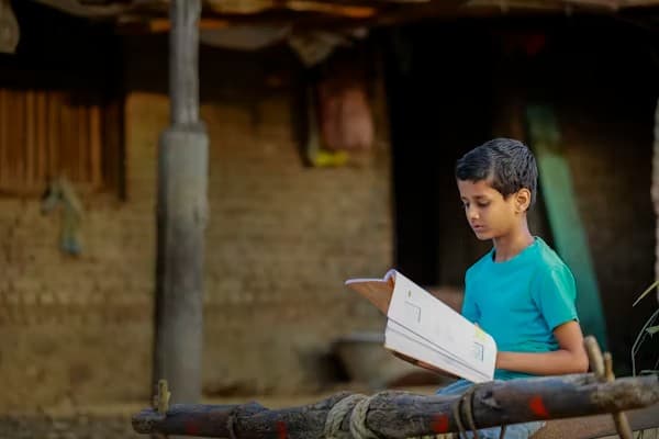 A young boy reading a textbook on a charpai outside his rural home, the quiet hope at the heart of every Sneha Sammilana programme.